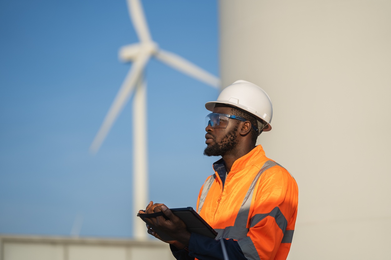 Field service engineer holding tablet standing near wind farm installation.