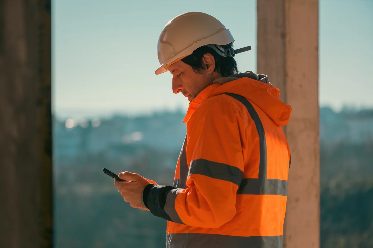 Field engineer in orange high-visibility coat holding tablet computer on skyscraper construction site.